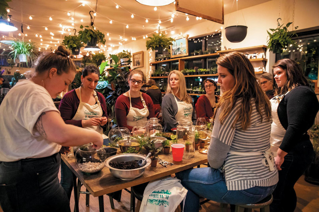 Women sit around a table at B. Willow in Baltimore to try their hand at terrarium making. In the background, fairy lights hang in rows on the ceiling and various plants hang from walls and sit on shelves. 