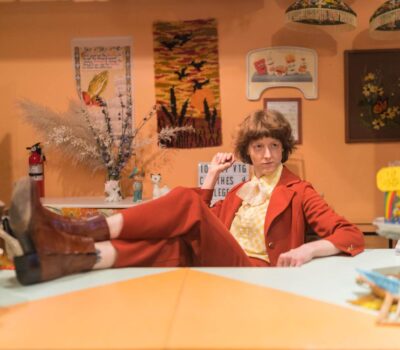 A person posing with their legs propped onto a table in a yellow and burnt orange vintage suit at Get Shredded Vintage in Baltimore.