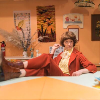 A person posing with their legs propped onto a table in a yellow and burnt orange vintage suit at Get Shredded Vintage in Baltimore.