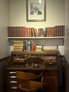 A recreation of an office featuring a wooden desk. A shelf of books sits over the desk, and a picture of Frederick Douglass hangs on the wall. 