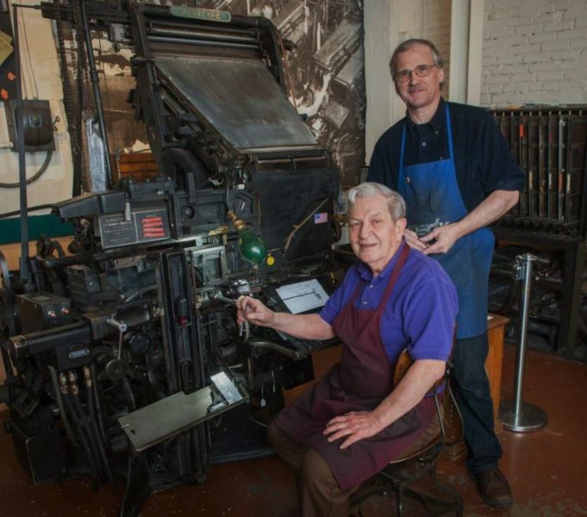 Two men in aprons posing by linotype press.