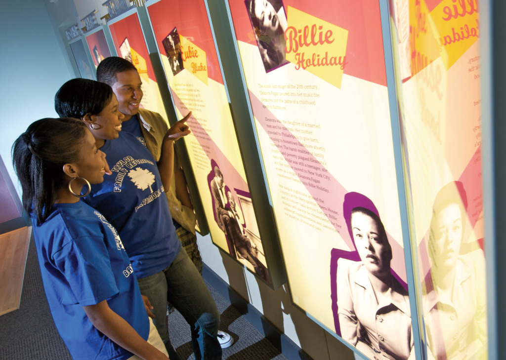 Two women and one man point at a description of Eubie Blake at the The Reginald F. Lewis Museum of Maryland African American History & Culture