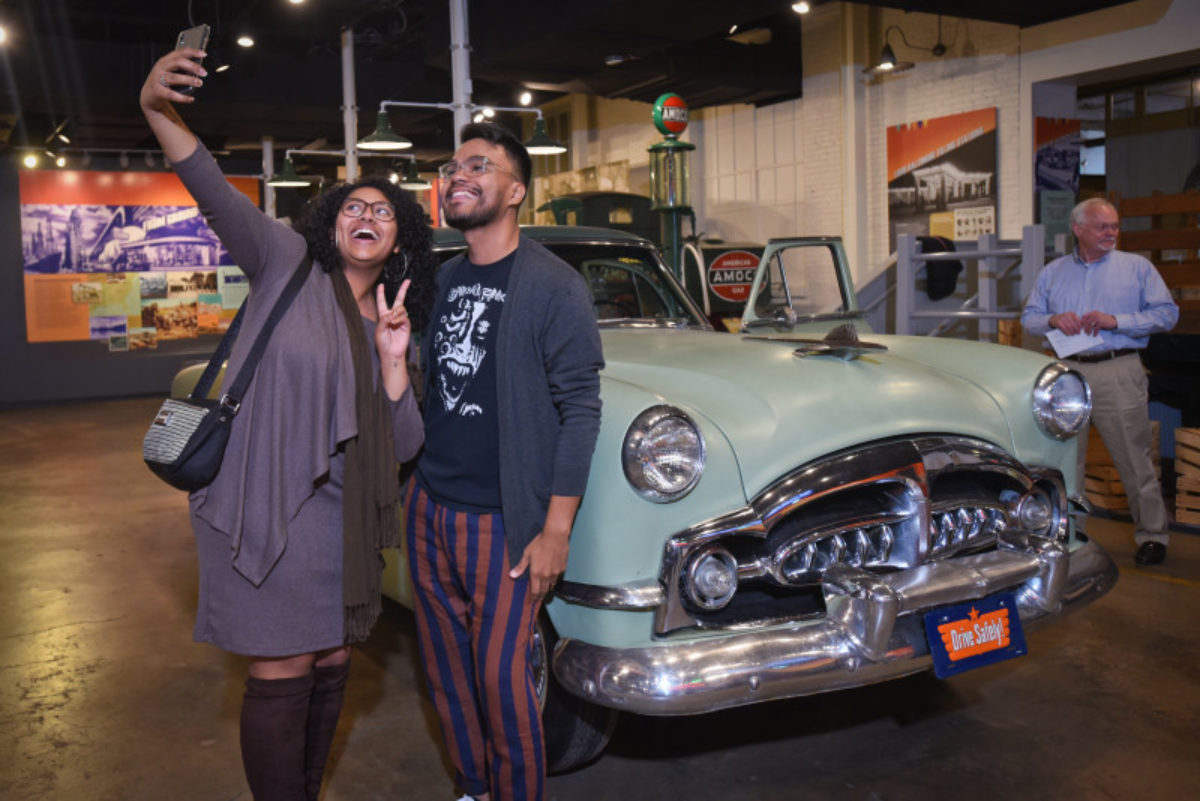 A man and woman take a selfie in front of a vintage car