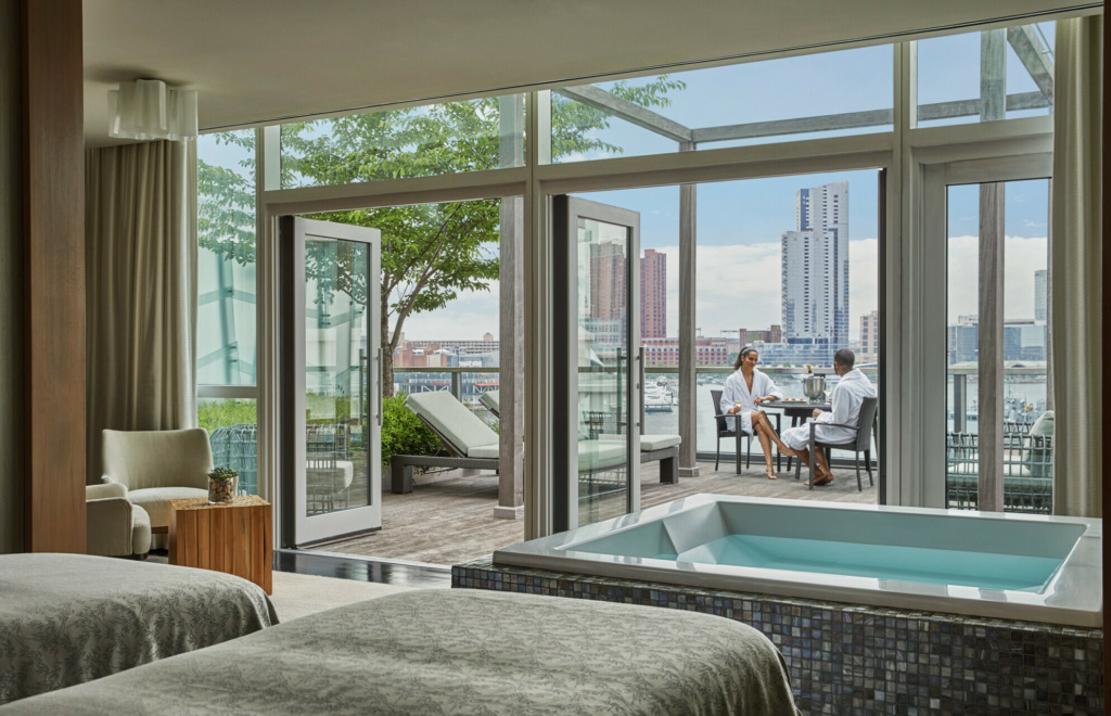 Wide shot of a waterfront Four Seasons Room. In the foreground are two beds and a hot tub. In the distance open balcony doors reveal a man and a woman enjoying breakfast on the patio. Both are wearing white towel robes. The Baltimore skyline is visible behind them.