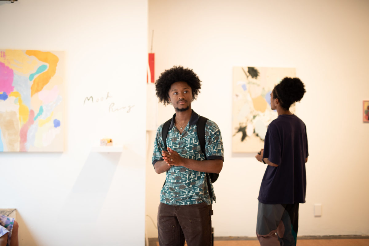 An African American man stands in the center of this photo. He is wearing a blue patterned shirt and a backpack. He is in a bright gallery and few paintings are visible in the background.