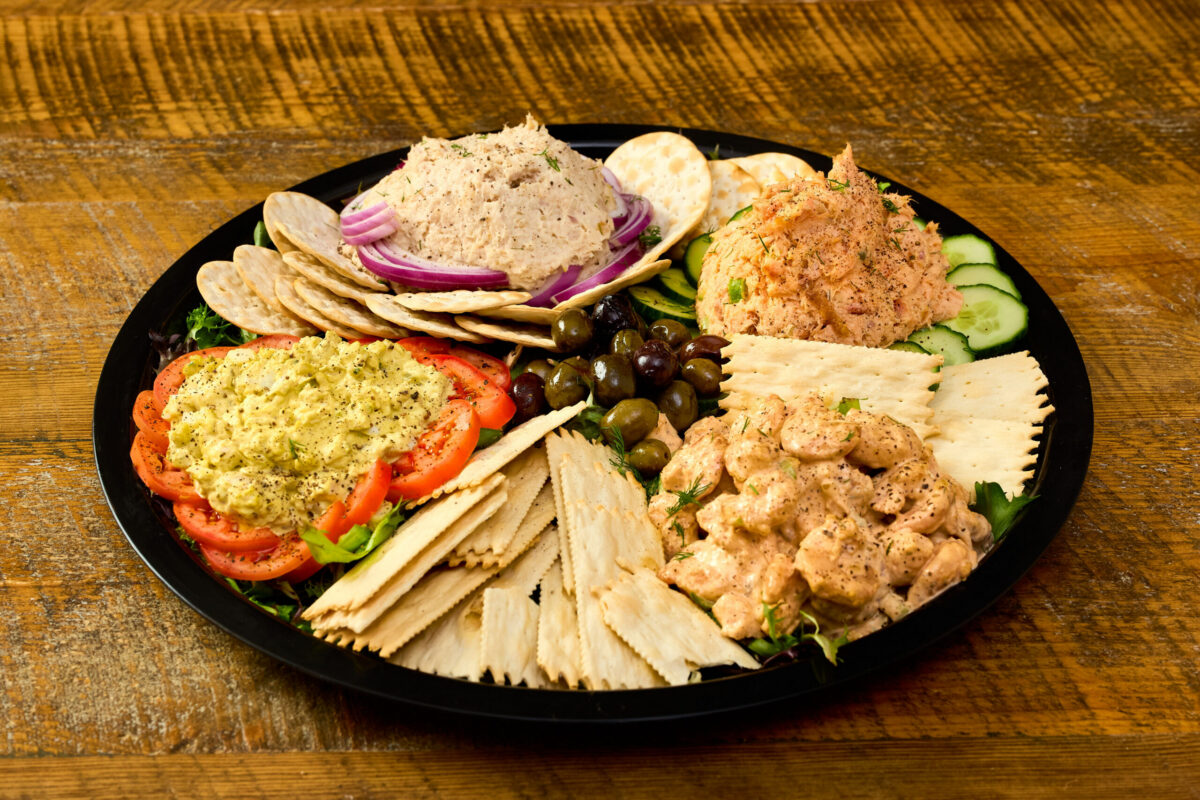 An assortment of smoked salad salad, white fish salad, shrimp salad, and smoked egg salad all displayed beautifully with sliced cucumbers, tomatoes, crackers and bagel chips.