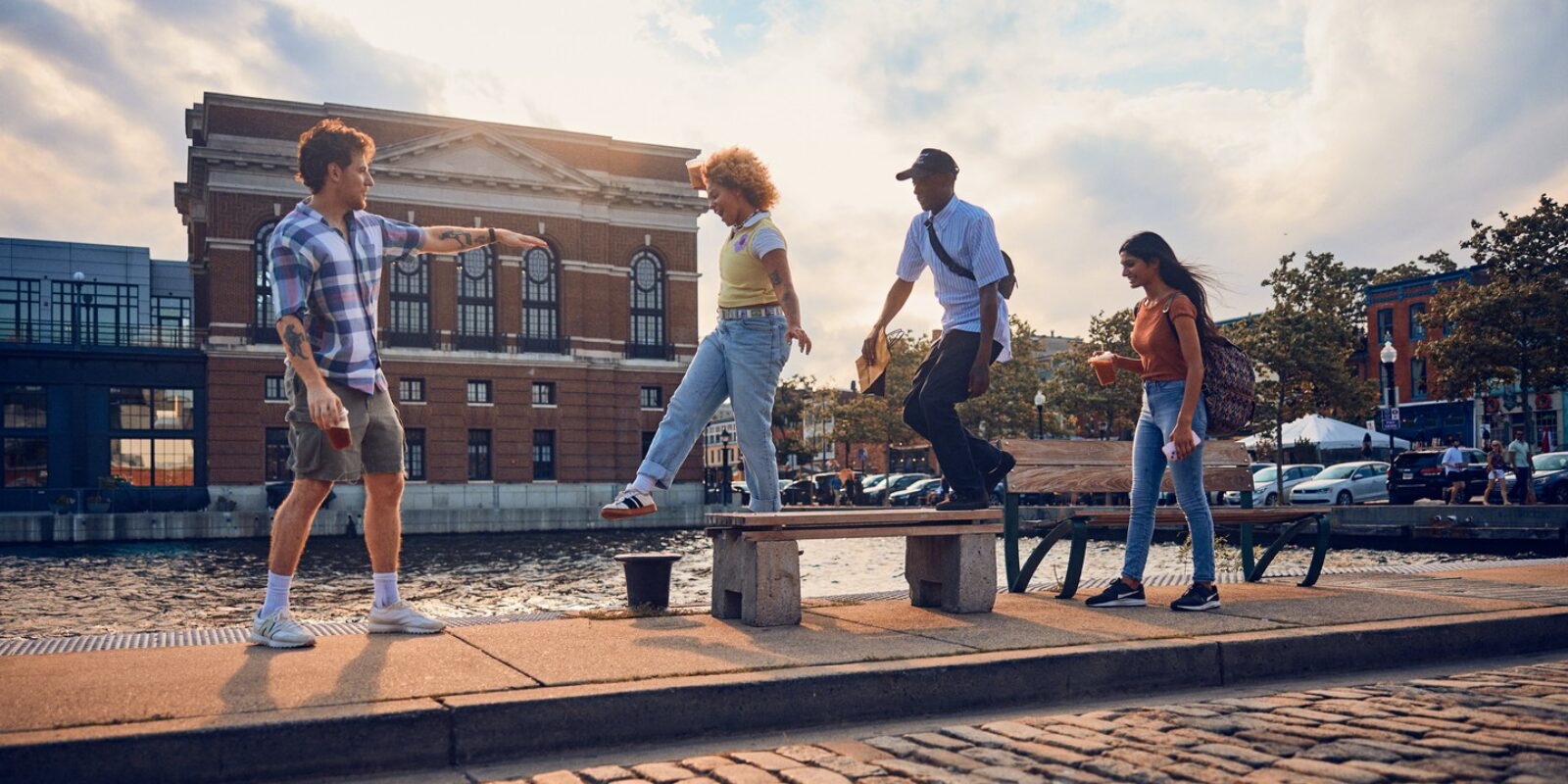 Four people walking along the sidewalk in Fell's Point. Two are standing on a bench.