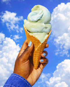 A dark-skinned hand with blue nail polish holds an ice cream cone against a blue sky background